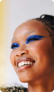 A person with a beaming smile, wearing dramatic blue glitter eyeshadow that extends into winged eyeliner.